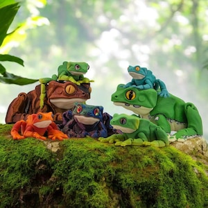 May include: A group of brightly coloured frog ornaments in different sizes and poses. The frogs are in shades of green, orange, brown, and blue, with detailed eyes and textures. They are arranged on a moss-covered surface, creating a whimsical scene.