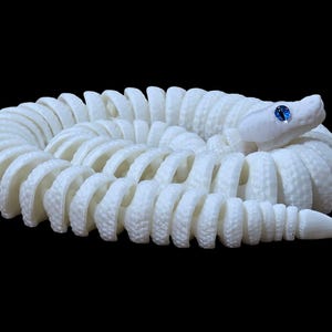May include: A coiled white snake sculpture with a textured surface. The snake has a detailed design and a blue gem for an eye. The sculpture is set against a black background.