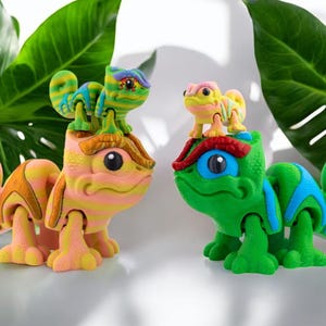 Può includere: Tre figurine di camaleonte colorate, un grande camaleonte verde con un camaleonte giallo più piccolo sulla testa, e un camaleonte rosa più piccolo con un camaleonte verde sulla testa.