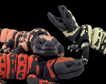 Pode incluir: Três figuras de lagarto de brinquedo, uma com um padrão preto, branco e amarelo, outra com um padrão preto, rosa e branco, e outra com um padrão preto, vermelho e branco.