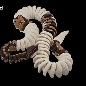 May include: A coiled, decorative snake sculpture with a segmented body. The snake features alternating sections of white and brown with a textured pattern. The head is brown with purple eyes. The word "Pied" is in the upper left corner.