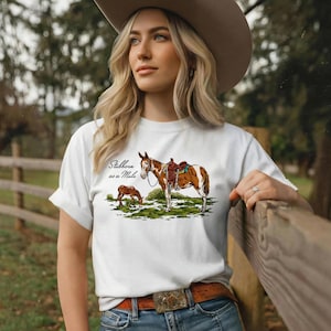 May include: White t-shirt featuring a graphic of a brown and white horse with a saddle, and a smaller brown horse grazing. The text "Stubborn as a Mule" is above the horse graphic. The person is wearing a brown belt and a cowboy hat.