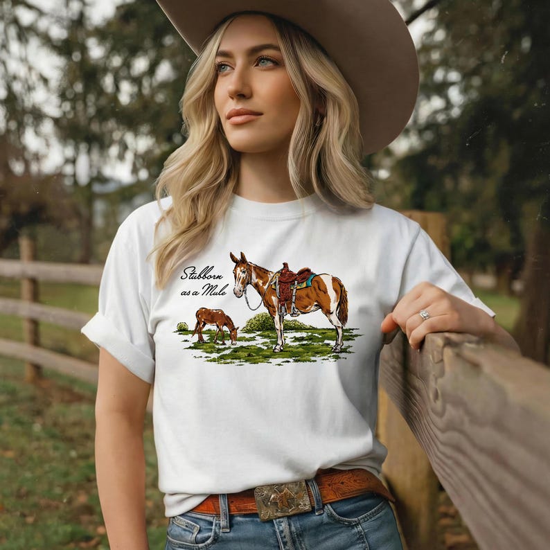May include: White t-shirt featuring a graphic of a brown and white horse with a saddle, and a smaller brown horse grazing. The text "Stubborn as a Mule" is above the horse graphic. The person is wearing a brown belt and a cowboy hat.