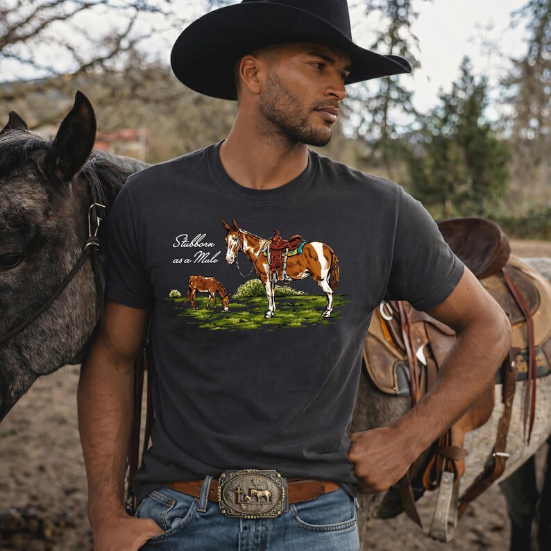 May include: Dark gray t-shirt featuring a graphic of a mule with saddle and a smaller mule, with the text "Stubborn as a Mule". The shirt is worn by a person wearing a cowboy hat and belt buckle.
