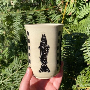 May include: A cream-colored ceramic tumbler featuring a black graphic print of a fish centered on the front. The sides of the tumbler are decorated with a vertical black and cream checkered pattern. The vessel has a smooth, cylindrical shape.