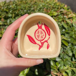 May include: A cream-colored ceramic wall pocket with a rounded arch shape and a small hole at the top for hanging. The center features a bright red graphic illustration of a halved pomegranate with leaves and a stem on a branch.