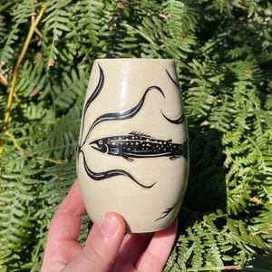 May include: A cream-colored ceramic tumbler featuring a black hand-drawn illustration of a fish swimming among flowing aquatic plants. The vessel has a smooth, rounded silhouette and a tapered opening, showcasing a clean, artistic design.