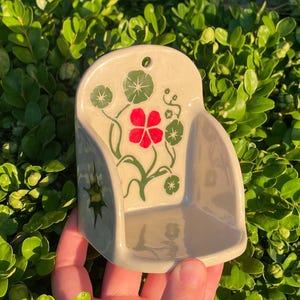 May include: A cream-colored ceramic wall pocket featuring a hand-painted design of green leaves and a single bright red flower. The piece has a curved back with a small hole for hanging and a smooth, glossy finish on the front surface.