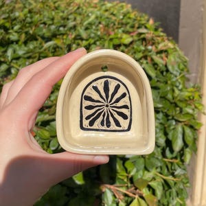 May include: A cream-colored ceramic wall pocket with an arched top and a small hole for hanging. The interior features a black hand-painted graphic of a stylized sunburst or flower design centered within an arched frame.