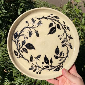 May include: A round, cream-colored ceramic plate featuring a black, hand-painted illustration of a blackberry vine. The vine curves around the perimeter of the plate, showcasing detailed leaves, thorny stems, and clusters of small, textured blackberries.