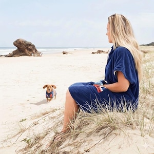 Op de afbeelding: Een marineblauwe poncho met capuchon, korte mouwen en een voorzak met een rood, wit en blauw gestreept patroon. Een kleine bruine teckel staat in de buurt en draagt een bijpassende trui met een rood, wit en blauw gestreept patroon.