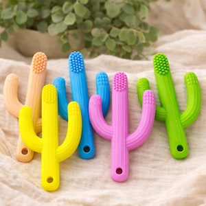 Könnte beinhalten: Eine Sammlung farbenfroher, kaktusförmiger Beißringe für Babys in Gelb, Orange, Blau, Rosa und Grün. Jeder Beißring hat eine strukturierte Oberfläche und ein Loch an der Basis. Die Beißringe liegen auf einem cremefarbenen Stoff.