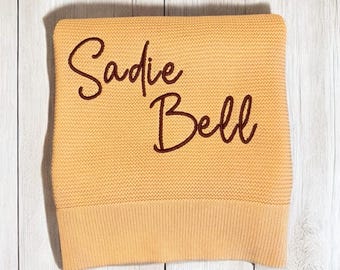 May include: A folded, light orange knitted blanket with the name "Sadie Bell" embroidered in a dark brown script. The blanket has a ribbed border and is displayed on a light wood-grain surface.