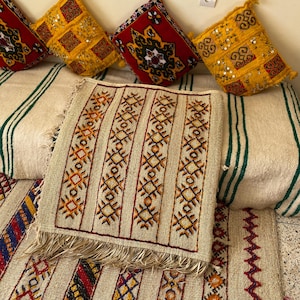 Puede incluir: Una colección de cojines y textiles decorativos con un diseño tradicional. Los cojines son rojos y amarillos con patrones y adornos intrincados. Los textiles presentan patrones geométricos en beige, naranja y rojo, con un borde con flecos. Los artículos están dispuestos sobre un fondo a rayas.