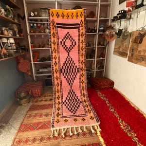 Op de afbeelding: Een roze en zwart geometrisch patroon tapijt met een gele rand en witte franjes. Het tapijt wordt verticaal weergegeven, waarbij het ontwerp wordt getoond. Het tapijt maakt deel uit van een collectie tapijten.