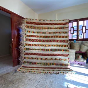 Puede incluir: Una alfombra beige y crema con un patrón geométrico de diamantes y rayas rojos, amarillos y verdes. La alfombra tiene un borde con flecos y se exhibe en interiores.