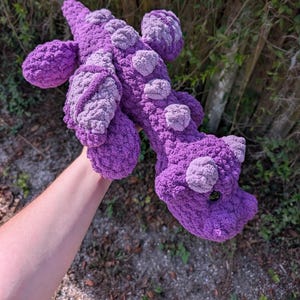 May include: A handmade, purple crochet dragon plushie with lighter purple accents. The dragon has a textured, bumpy design and is held up against a background of green foliage.