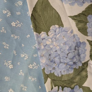 May include: A fabric featuring two distinct patterns: one side displays a light blue background with a small white floral print, while the other side shows a white background with large blue hydrangea clusters and dark green leaves.