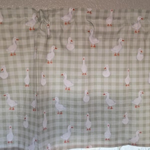 May include: A valance featuring a repeating pattern of white geese with orange feet and beaks on a light green and white checkered background. The valance is made of fabric and is designed to be hung above a window.