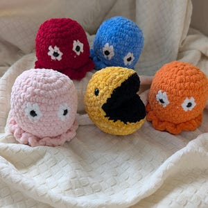 May include: Five colorful crocheted plush toys in the shape of octopuses and Pac-Man. The toys are red, blue, pink, orange, and yellow. Each octopus has white flower-shaped eyes. The Pac-Man has a black mouth.