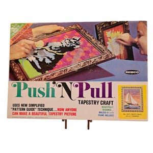 May include: A rectangular box for a Push 'N Pull tapestry craft kit. The box features illustrations of a cat and a sailboat, with text including 'Push 'N Pull', 'Tapestry Craft', and 'Molded 8 1/2 x 11 frame included' printed on the front.