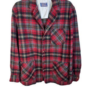 Könnte beinhalten: Ein rot-schwarz-grau karierter Blazer mit zwei Vordertaschen und einer Brusttasche. Der Blazer hat ein fallendes Revers und einen Zwei-Knopf-Verschluss. Ein kleines Etikett ist auf der Innenseite des Kragens sichtbar. Der Blazer ist aus dickem, gewebtem Stoff.