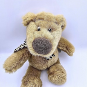 Puede incluir: Un oso de peluche con un cuerpo marrón y peludo, una gran nariz marrón y pequeños ojos negros. El oso tiene una bufanda a cuadros en blanco y negro. Un peluche.