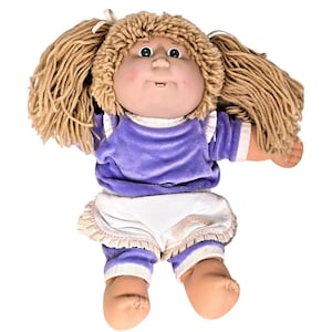 Może przedstawiać: Zabytkowa lalka Cabbage Patch Kids z blond włosami z przędzy w kucykach. Lalka ma na sobie fioletowy aksamitny top, białe szorty z marszczonymi krawędziami oraz pasujące fioletowo-białe legginsy w paski. Lalka ma różowe policzki i uśmiechniętą minę.