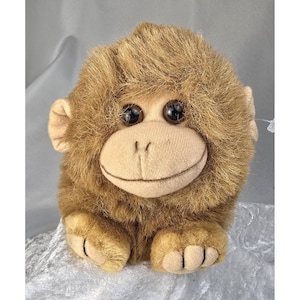 May include: A plush monkey toy with brown fur, large dark eyes, and a smiling face. The toy has a light brown face, ears, and feet. The monkey is sitting upright, with its arms at its sides. A tag is attached to the toy.