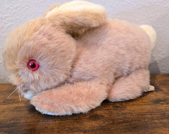 May include: A plush rabbit toy with soft, light tan fur and bright red eyes. The stuffed animal is in a resting position with its ears folded back against its head and a small white tail visible at the rear. It has thin white whiskers.