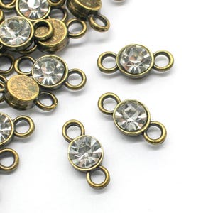 May include: Close-up of antique brass-toned connectors with clear, round rhinestones. The connectors have a circular rhinestone in the center and two small loops on each side. A pile of connectors is on the left side of the image.