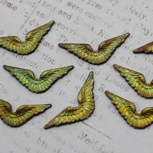 May include: Eight small, gold-toned metal angel wings with a green iridescent finish. The wings are arranged in a random pattern on a white surface with printed text.