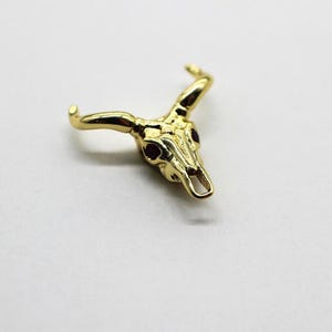 May include: A gold-coloured bull skull pendant with dark red accents in the eye sockets. The pendant has detailed horns and a textured surface, set against a plain white background. This jewellery piece is likely a charm or a small decorative item.