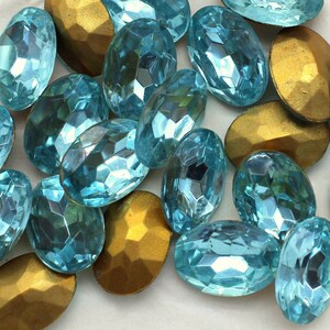 May include: A collection of oval-shaped, turquoise blue glass rhinestones with a faceted finish. The rhinestones are scattered on a white surface with several gold-colored metal pieces.
