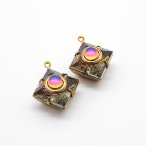 Könnte beinhalten: Zwei goldfarbene quadratische Anhänger mit irisierenden Glascabochons. Die Cabochons haben einen Farbverlauf von Pink, Lila und Blau. Die Anhänger haben eine strukturierte, metallische Oberfläche.