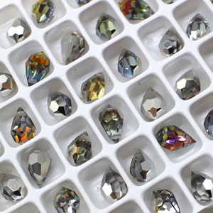 May include: A white tray filled with numerous faceted, teardrop-shaped rhinestones. The rhinestones vary in color, including silver, iridescent, and shades of gold, orange, and purple. The tray's grid pattern neatly organizes the gems.