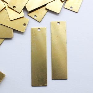 Könnte beinhalten: Eine Sammlung rechteckiger Messinganhänger mit einem kleinen Loch oben. Die Anhänger haben eine warme Goldfarbe und variieren leicht in der Größe. Einige Anhänger sind verstreut, andere sind ordentlich angeordnet.