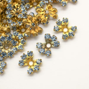 May include: A pile of gold-toned metal flower-shaped embellishments with light blue rhinestones.