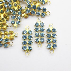 May include: A close-up of several gold-toned metal embellishments with light blue rhinestones. The embellishments are shaped like a rectangle with a loop at the top.
