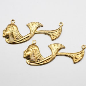 May include: Two gold-toned metal charms featuring an Egyptian pharaoh's head with a headdress and a lotus flower design.