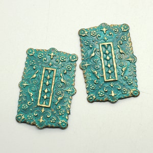 Könnte beinhalten: Zwei tealgrüne, metallene Dekorverzierungen mit goldenen Akzenten. Die Verzierungen sind rechteckig mit einem verzierten Design und einem goldenen vertikalen Rechteck in der Mitte.