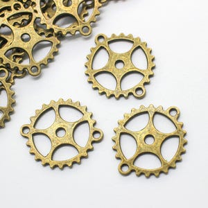 May include: Several antique brass-colored gear charms with a steampunk aesthetic. The gears have a circular design with multiple spokes and small holes, perfect for jewelry making or crafting projects. The charms are on a white background.