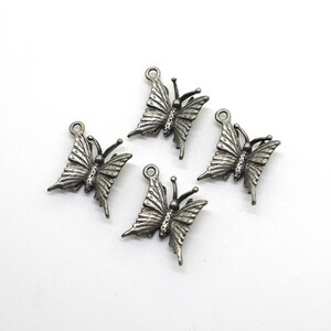 Könnte beinhalten: Vier kleine, silberfarbene Schmetterlingsanhänger mit detaillierten Flügelmustern und einer Schlaufe oben zur Befestigung an Schmuck oder anderen Bastelarbeiten. Die Schmetterlinge sind auf weißem Hintergrund angeordnet.