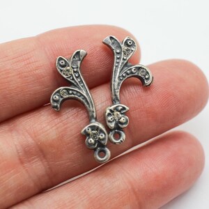 Könnte beinhalten: Zwei silberfarbene, verzierte Metallanhänger. Jeder Anhänger hat ein stilisiertes Blumendesign mit gebogenen Stielen, kleinen kreisförmigen Akzenten und einer Schlaufe unten. Die Anhänger sind wahrscheinlich für die Schmuckherstellung oder Verzierung gedacht.