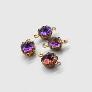May include: Four iridescent, faceted glass beads with a metallic gold-coloured setting and small loops for stringing. The beads have a purple and red colour shift. They are set against a white background.