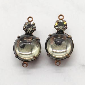May include: Two vintage-style earrings. Each earring features a clear, round glass orb with a reflective interior, set in a bronze-colored metal frame. A smaller, faceted crystal tops each orb. The earrings are displayed on a white marble surface.