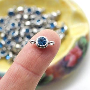 May include: A small, round, silver-colored stud earring with a dark blue gemstone center. The earring is held between two fingers, with a blurred background of a yellow and floral-decorated ashtray filled with similar blue gemstones.