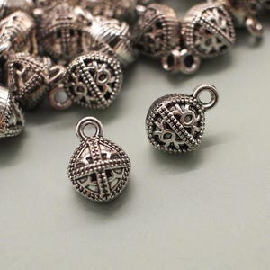 May include: A collection of small, silver-colored metal charms with intricate openwork designs. The charms are spherical with a loop at the top for attaching to jewelry. The background is a light gray surface.