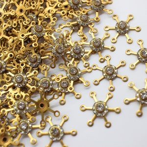 May include: A close-up of a pile of gold-toned metal snowflake charms with clear rhinestones in the center. The charms are scattered on a white surface.