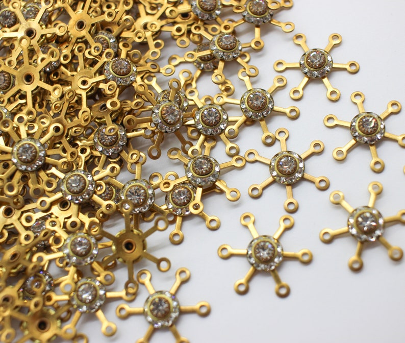 May include: A close-up of a pile of gold-toned metal snowflake charms with clear rhinestones in the center. The charms are scattered on a white surface.